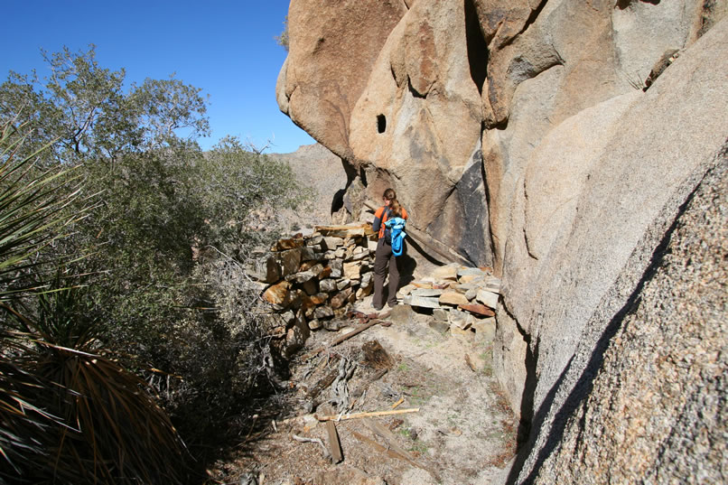 Perched on the edge of the cliff a short distance from the shaft is the old blacksmith work area.  Notice the remains of a stacked stone forge behind Niki and the rectangular opening chiseled into the cliff above her head.  We wonder if a beam had once been set into the opening.